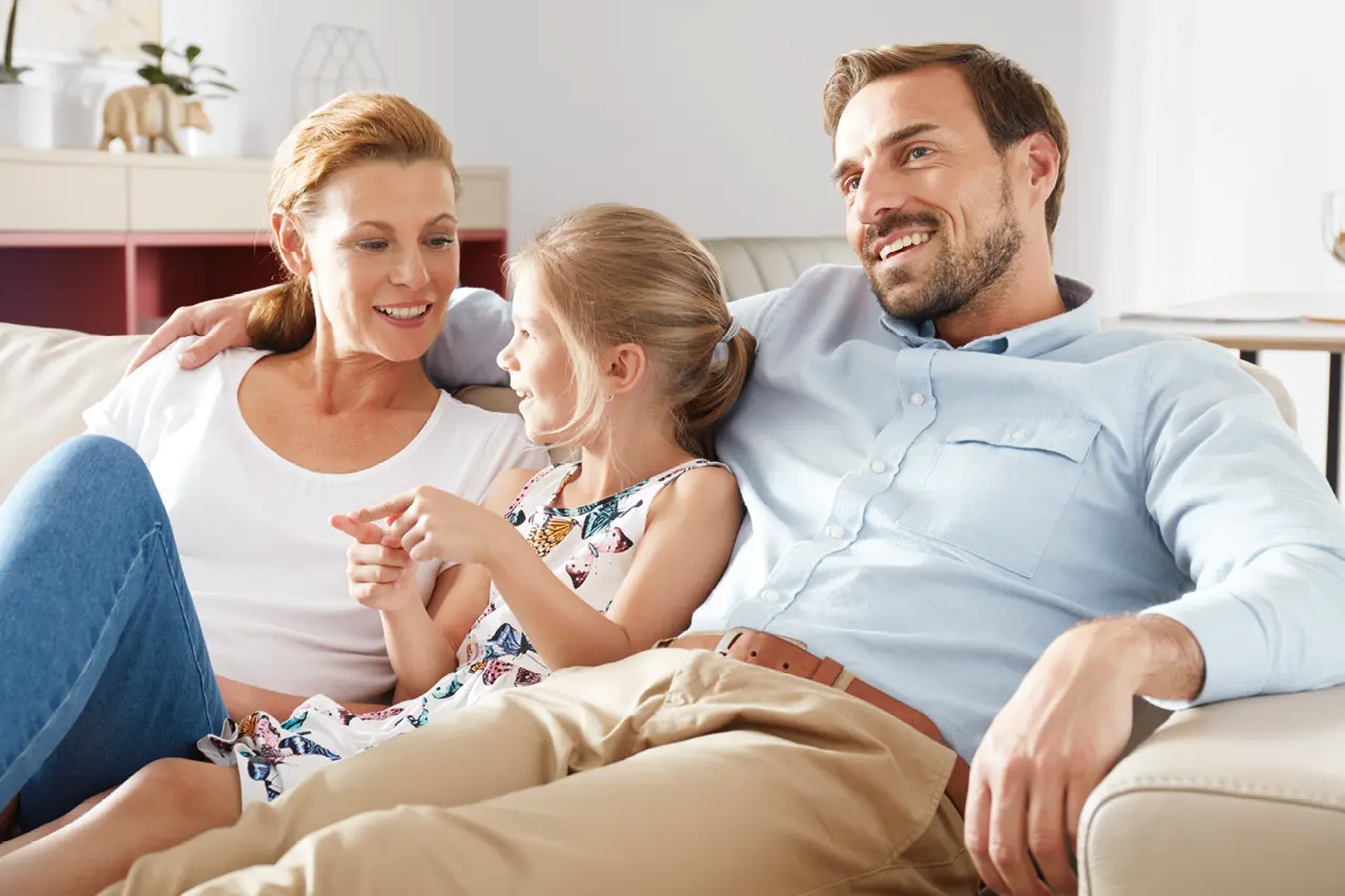 Glückliche Familie mit Mutter, Vater und Kind sitzt entspannt auf einem Sofa im Wohnzimmer und unterhält sich, helle Wohnmöbel und freundliche Atmosphäre stehen für modernes Familienleben.